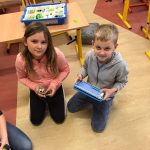 Dziewczynka i chłopiec klęczą na podłodze. Chłopiec trzyma w ręce tablet, dziewczynka klocki