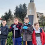 Czterech chłopców z narzędziami do sprzątania stoi pod pomnikiem. Na postumencie krzyż, tablica oraz figura orła
