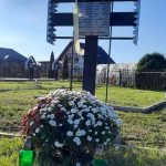 Krzyż na cmentarzu wojskowym, na nim tablica z nazwiskami poległych, pod nim duży bukiet kwiatów i dwa znicze