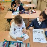 Dwóch chłopców siedzi w ławce i koloruje obrazek. Za nimi siedzi jeden chłopiec, w ręce trzyma kredki, przygląda się obrazkowi