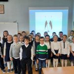 Grupa uczniów stoi w sali w sali lekcyjnej na tle wyświetlonych na tablicy multimedialnej flag polskich