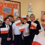 Sześciu chłopców ubranych na galowo stoi pod gazetką w sali lekcyjnej. W rękach trzymają flagi Polski. Obok stoi nauczycielka. Na przodzie widać dziewczynkę wykonującą zdjęcie