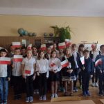 Uczniowie w sali lekcyjnej stoją w grupie. Każdy z nich w prawej dłoni trzyma biało - czerwoną flagę