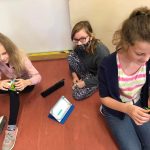 Trzy dziewczynki kucają na podłodze. Dwie trzmają w rękach samochody z klocków, trzecia patrzy w obiektyw. Między nimi stoją dwa tablety