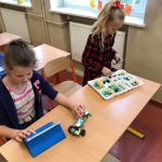 Dwie dziewczynki siedzą w ławce, budują robota z klocków lego. Jedna trzyma w ręce tablet