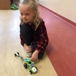 Dziewczynka kuca na podłodze w sali lekcyjnej, bawi się samochodem z klocków