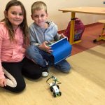 Dziewczynka i chłopiec w sali lekcyjnej klęczą na podłodze, przed nimi jeżdżący robot - samochód. Chłopiec trzyma w ręce tablet