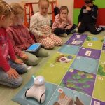 Na kolorowej macie stoi robot. Wokół maty siedzą dzieci, jedno z nich trzyma tablet