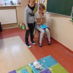 Dziewczynka siedzi na krześle, programuje na tablecie. Przy niej stoi chłopiec, który się przygląda