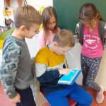 Chłopiec siedzi na krzesełku w sali lekcyjnej, programuje na tablecie. Nad nim stoją dwie dziewczynki i jeden chłopiec