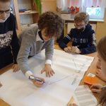 Przy stoliku siedzi jedna dziewczynka i dwóch chłopców. Druga dziewczynka pochyla się nad stolikiem zapisując pomysły grupy na dużej kartce papieru