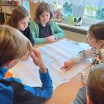 Trzy dziwczyny i jeden chłopiec siedzą przy ławce w sali. Na ławce rozłożona jest duża kartka papieru, na której jedna z dziewcząt wskazuje palcem