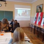 Uczniowie siedzą w sali lekcyjnej, oglądają wyświetlany na projektorze film. Z prawej strony dekoracja okolicznościowa