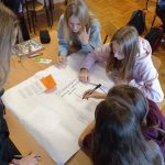 Cztery dziewczynki pracują w grupie przy stoliku. Na stoliku rozłożona jest karta papieru, na której jedna z dziewcząt rysuje linię