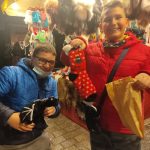 Roześmiany chłopiec w czerwonej kurtcetrzyma czerwoną skarpetę. Obok niego uśmiechnięty drugi chłopiec, za nimi stragan z pamiątkami