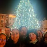 Uśmiechnięte twarze siedmiu dziewczynek i jednego chłopca. Za nimi duża, rozświetkona choinka oraz budynki