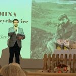 Chłopiec w szarej marynarce i czarnych spodniach stoi na scenie, w ręce trzyma mikrofon, recytuje wiersz.W tle zdjęcie poety