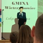Chłopiec w czarnym garniturze stoi na scenie i recytuje wiersz. W tle napis Gmina Wietrzychowice