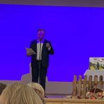 Mężczyzna trzyma w ręce kartki papieru, stoi na scenie, mówi do mikrofonu