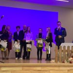 Grupa uczniów na scenie, obok mężczyzna w okularach trzymający w ręku mikrofon. Przed mężczyzną niski płotek oraz wysypane z koszyka jabłka