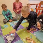 Chłopiec głaszcze robota stojącego na macie. W tle dwie dziewczynki obserwują sytuację