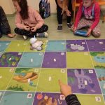 kolorowa mata, na niej robot. przed matą dwie dziewczynki, jedna trzyma tablet, druga coś do niej mówi. Trzecia dziewczynka siedzi po turecku odwrócona w stronę innego dziecka