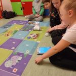 Czworo dzieci siedzi na podłodze, przed nimi leży mata, na której jest robot. Dzieci zajmują się programowaniem