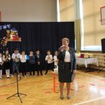 Uśmiechnięta kobieta stoi na sali gimnastycznej, mówi do mikrofonu. Za nią grupa dzieci oraz stolik z książkami