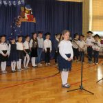 Dziewczynka ubrana w białą bluzkę i granatową spódnicę stoi przed mikrofonem. Za nią stoi grupa dzieci ubranych w biało - granatowe stroje