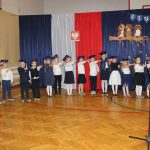 Grupa dzieci z klasy pierwszej wygłasza tekst ślubowania. Dzieci ubrane są na galowo, każde ma wyciągniętą prawą rękę w geście ślubowania