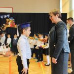 Pani dyrektor pasuje chłopca na ucznia szkoły trzymając na jego ramieniu drewniany, długi ołówek. W tle grupa dzieci na sali gimnastycznej