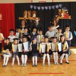 Uczennice klasy pierwszej siedzą na ławce, za nimi stoją uczniowie, w trzecim rzędzie wychowawczyni, pani dyrektor i wójt