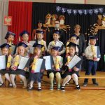 Grupa uczniów klasy pierwszej po ślubowaniu, za nimi dwie kobiety, między kobietami stoi mężczyzna. W tle granatowy materiał, na nim napis: ślubujemy, flaga biało-czerwona, godło