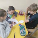 Dwie dziewczynki i jeden chłopiec siedzą przy żółtym stoliku i rozwiązują zadanie na kartce.