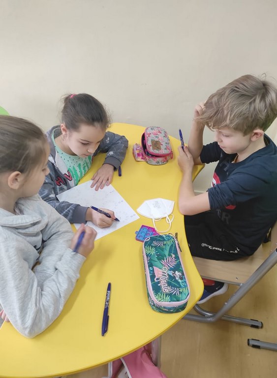 Dwie dziewczynki i jeden chłopiec siedzą przy żółtym stoliku i rozwiązują zadanie na kartce.