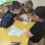 Dwóch chłopców i jedna dziewczynka siedzą przy żółtym stoliku i rozwiązują zadania na białej kartce.