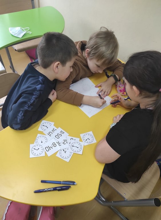 Dwóch chłopców i jedna dziewczynka siedzą przy żółtym stoliku i rozwiązują zadania na białej kartce.