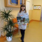 Dziewczynka w szarej sukience stoi na korytarzu szkolnym. Prezentuje pracę namalowaną farbami, przedstawiającą mężczyznę na koniu, w tle rzeka i las