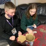 Dziewczynka i chłopiec siedzą na czarnej, skórzanej kanapie przy stoliku przykrytym serwetą. Na stoliku leżą wysypane z puszki pieniądze