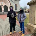 Dwaj chłopcy ubrani w ciepłe kurtki stoją w bramie ogrodzenia. Za nimi fragment kościoła