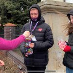 Chłopiec w czarnej kurtce z naklejonym serduszkiem WOŚP trzyma puszkę. Obok niego stoi dziewczynka, w ręku trzyma naklejki WOŚP. Przed nimi stoi kobieta w różowym polarze, która wrzuca datek do puszki chłopca