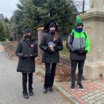 Dwie dziewczynki i jeden chłopiec stoją przed ogrodzeniem. Dziewczynka w środku trzyma puszkę WOŚP