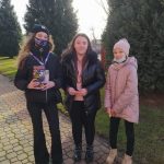 Trzy dziewczyny stoją na chodniku, jedna trzyma w rękach puszkę Wielkiej Orkiestry Świątecznej Pomocy