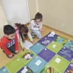 Dwie dziewczynki i jeden chłopiec siedzą na podłodze obok kolorowej maty i białego robota rozwiązując zadania na białej kartce papieru.