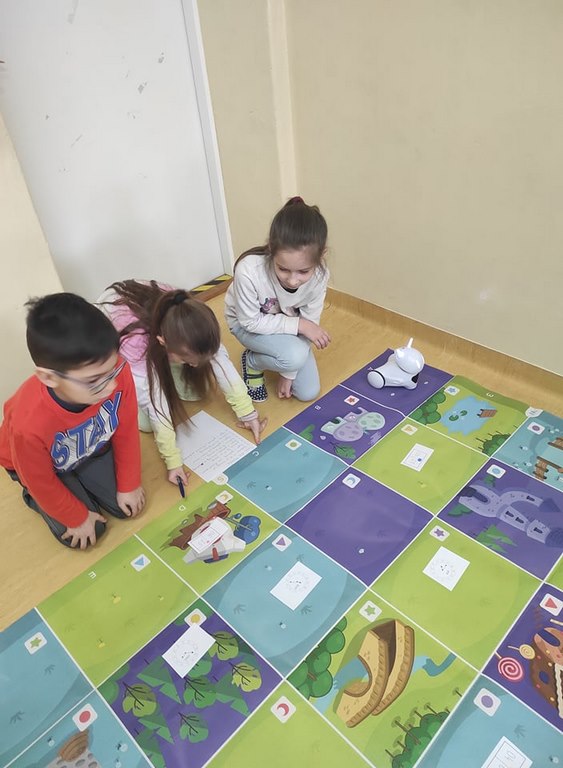 Dwie dziewczynki i jeden chłopiec siedzą na podłodze obok kolorowej maty i białego robota rozwiązując zadania na białej kartce papieru.