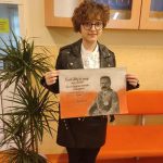 Dziewczyna w okularach stoi na korytarz szkolnym, w rękach trzyma dużą kartkę papieru z wizerunkiem Piłsudskiego na biało - czerwonym tle