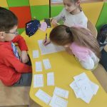 Dwie dziewczynki i jeden chłopiec siedzą przy żółtym stoliku, na którym rozrzucone są białe karteczki z godzinami i rozwiązują zadania na białej kartce papieru.