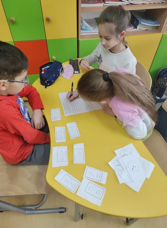 Dwie dziewczynki i jeden chłopiec siedzą przy żółtym stoliku, na którym rozrzucone są białe karteczki z godzinami i rozwiązują zadania na białej kartce papieru.