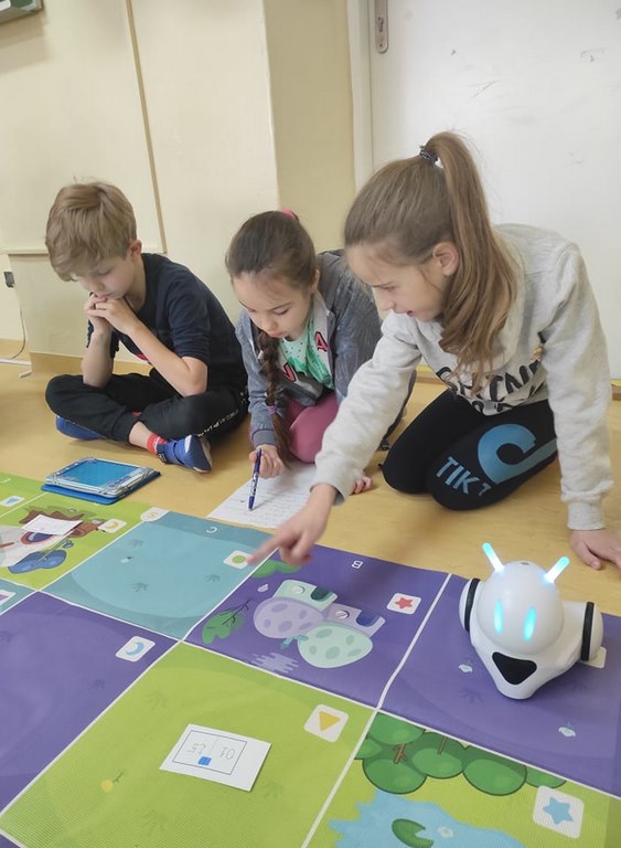 Troje dzieci siedzi na podłodze obok kolorowej maty, na której stoi biały robot.