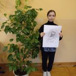 Dziewczynka w czarnych spodniach i czarnym golfie stoi obok dużego kwiatka, w rękach trzyma portret mężczyzny wykonany ołówkiem